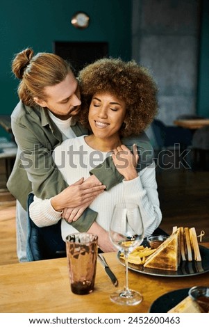 An African American woman and a man share a heartfelt hug at a table in a modern cafe, expressing love and connection. Royalty-Free Stock Photo #2452600463