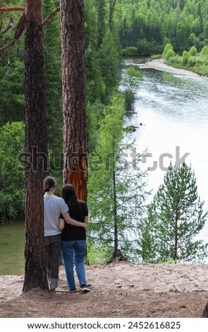 A couple of tourists admires beautiful river and green forest from a high steep bank on sunny day. Summer hikes and travels. River Ekhe-Ukhgun, foothill Tunka valley, Buryatia, Baikal region, Siberia Royalty-Free Stock Photo #2452616825