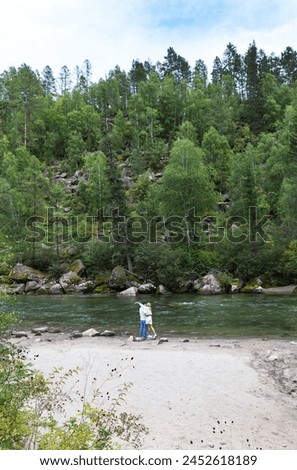 A tourist couple in love kisses on sandy beach of beautiful river against backdrop of green forest on vacation on sunny day. Summer trips and travels. River Ekhe-Ukhgun, Tunka, Buryatia, Baikal region Royalty-Free Stock Photo #2452618189