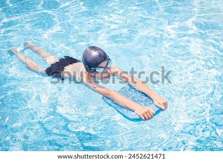 Active, happy child (boy) in cap and sport goggles doing water sport with swim board in the swimming pool. Healthy lifestyle. Kid enjoying water.