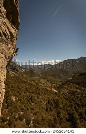 A person is climbing a rock face in the mountains. The sky is clear and the mountains are covered in trees