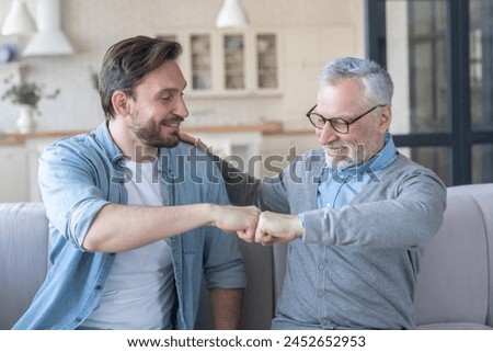Old elderly senior father together with his young adult son doing fists gesture, giving fists together at home showing support and love. Happy father`s day! I love you, dad!