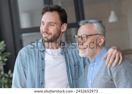 Young adult caucasian son hugging supporting his old elderly senior father at home indoors looking at the window in future. Happy father`s day! Care and love concept. I love you, dad!
