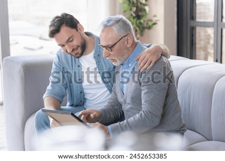 Caring loving adult son hugging embracing his old elderly senior father while he is showing his family photograph photo at home, telling stories of his youth. Happy father`s day! I love you, dad!