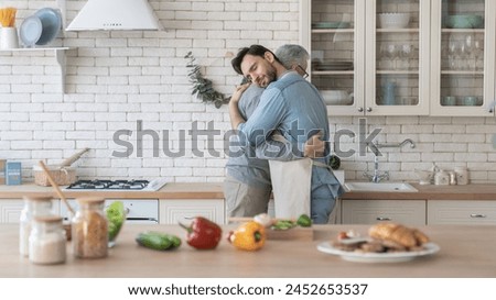 Side view image of a young adult caucasian son hugging embracing showing his care and love to elderly old senior father while cooking meal, food in the kitchen. Happy father`s day! I love you, dad!