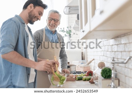 Cheerful adult son helping his senior old father in cooking, preparing family meal, dinner, lunch. Parents visiting. Happy father`s day! I love you, dad! Men cooking together