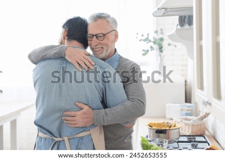 Cheerful loving caring old elderly senior father embracing hugging his adult caucasian son in the kitchen while cooking lunch, dinner, preparing meal together. Happy father`s day! I love you, dad!