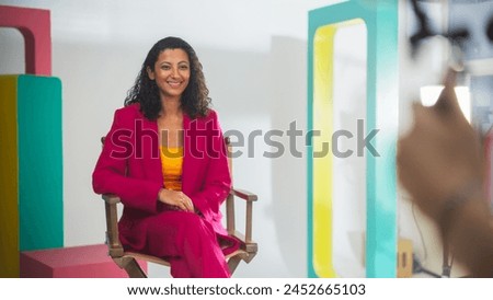 Young Middle Eastern Female Host In Vibrant Pink Blazer And Yellow Top, Seated On Director's Chair On Colorful Film Set, Smiling At Camera, With Blurred Videographer In Foreground. Royalty-Free Stock Photo #2452665103