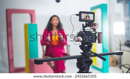 Vibrant Video Production Scene With A Black Female Host In A Pink Suit Engaging In A Lively Discussion, Captured On A Professional Camera Setup Against A Colorful, Modern Backdrop.