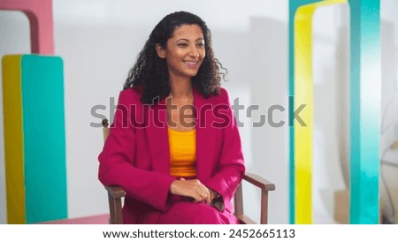 Young Black Female Host In Vibrant Pink Suit And Yellow Top, Smiling Engagingly On Colorful Set, Exemplifies Modern Videography And Dynamic Storytelling In Media Production.
