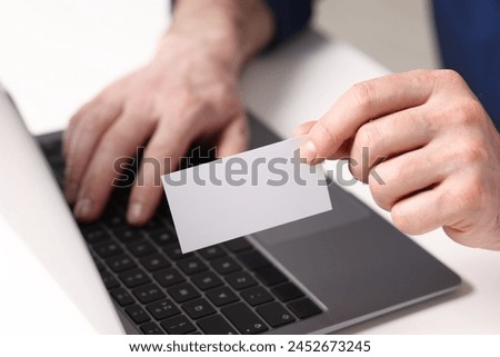Man with laptop holding blank business card at white table, closeup