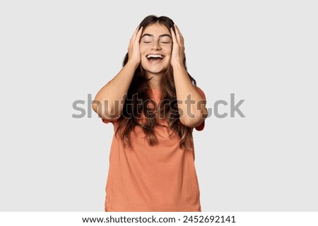 Young Caucasian woman in studio setting laughs joyfully keeping hands on head. Happiness concept.