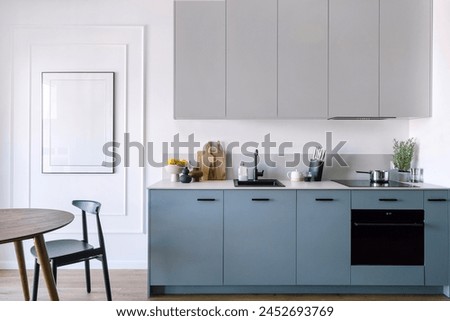 Kitchen with modern interior and household appliances, electric oven, induction hob, sink with water tap, blue cupboard, dining table with chair and empty picture on wall.