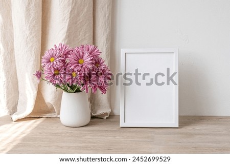 White empty picture frame mockup, vase with pink daisy flowers bouquet on wooden table, empty white wall background, beige linen curtain with natural sunlight shadows, aesthetic home interior.