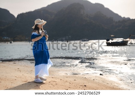 Woman enjoying the beach in summer, wearing a hat, smiling, with a  holding a camera taking pictures of the seascape