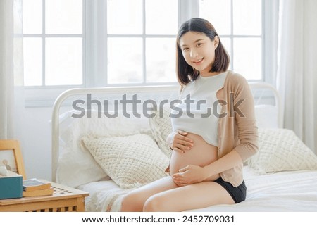 Young Asian happy pregnant woman is sitting and relaxing on bed and and touching her belly. Family plan, pregnancy, motherhood, people and expectation concept