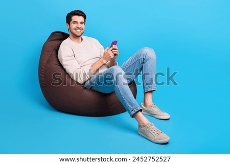 Full size photo of nice young man sit bag use smart phone wear beige sweatshirt isolated on blue color background