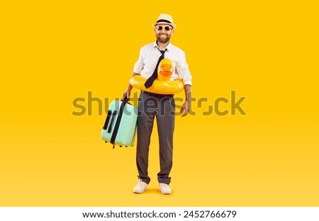 Office worker in formal wear isolated on a orange background, suitcase in hand and an inflatable ring nearby. Captures the essence of a well deserved holiday escape and spirit of summer vacation. Royalty-Free Stock Photo #2452766679