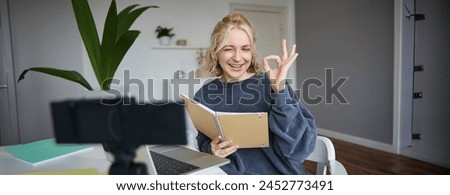 Portrait of cute blond girl with notebook in hands, shows okay sign, records video, social media content, blogging from her room.