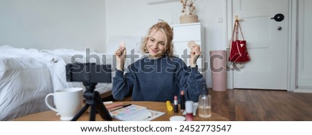 Image of young woman, vlogger records video on digital camera, shows beauty products, recommending makeup for audience on social media, sits on front in a room.