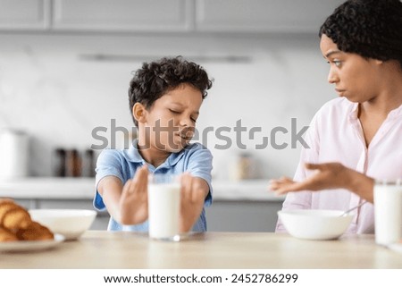 The image illustrates African American boy refusing a glass of milk with a grimace, showcasing a child's aversion or dietary choices in a family kitchen Royalty-Free Stock Photo #2452786299