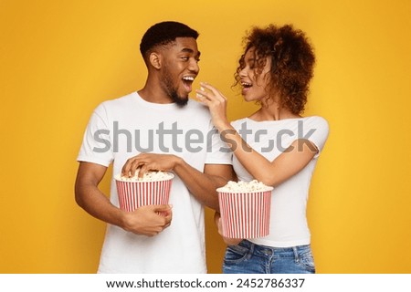 Black millennial couple in love enjoying popcorn together, feeding each other, orange studio background Royalty-Free Stock Photo #2452786337