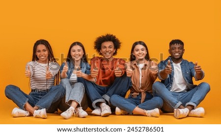 A diverse team of five friends give thumbs up while sitting, displaying approval and friendship in a multiethnic, multiracial context, isolated on a yellow background Royalty-Free Stock Photo #2452786501