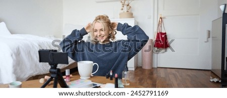 Portrait of cute blond girl, lifestyle blogger sits on floor with video camera and stabiliser, shows how to do hairstyle and makeup, does casual vlogging for social media, sits in room.