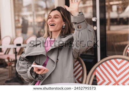 Beautiful young woman looking at camera hold mobile phone sitting in cafe and wave her hand, sign hi, hello. Blonde wearing trench coat and pink sweater sitting on the chair in restaurant.