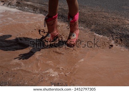A young girl is standing in a muddy puddle wearing red boots. Co