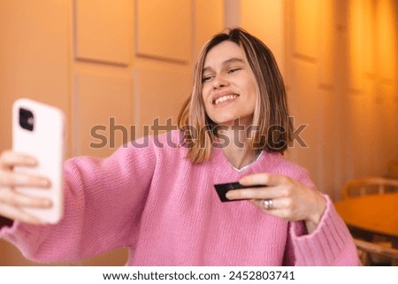 Image of a beautiful amazing happy excited young blonde short hair woman sitting in cafe take selfie by camera with plastic credit card. Girl wear pink sweater.