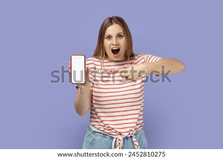 Portrait of amazed surprised blond woman wearing striped T-shirt holding cell phone with white empty display with copy space for promotional text. Indoor studio shot isolated on purple background.