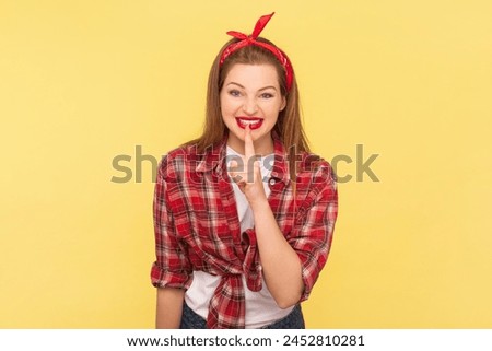 Portrait of positive cheerful woman keeps finger near red lips keeping secret, smiling with cunning look, wearing checkered shirt and headband. Indoor studio shot isolated on yellow background.