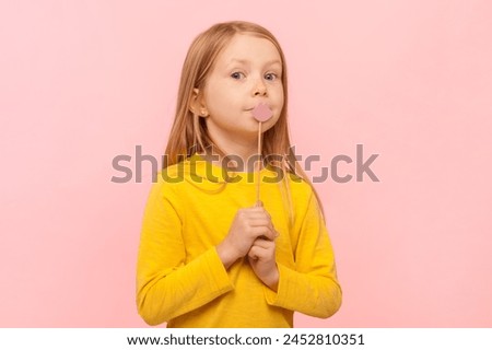 Portrait of funny cute blonde little girl standing with paper lips, party props, looking at camera, wearing yellow jumper. Indoor studio shot isolated on pink background.
