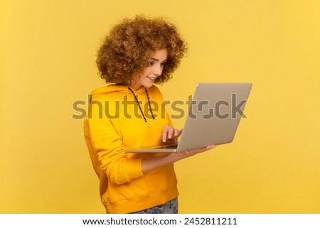 Side view portrait of smiling happy woman with Afro hairstyle holding laptop, typing on keyboard, searching information, wearing casual style hoodie. Indoor studio shot isolated on yellow background.