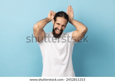Portrait of childish man with beard wearing white T-shirt showing bunny ears with hands on his head and looking at camera with childish toothy smile. Indoor studio shot isolated on blue background. Royalty-Free Stock Photo #2452811315