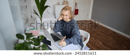 Lifestyle image of young woman writing down something in notebook, making notes, studying online, doing course in internet, listening to interesting information, using laptop.