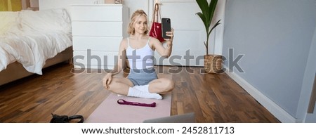Portrait of stylish young woman, sitting on rubber mat on floor at home, taking selfie while doing workout, yoga session, posting photo on social media.