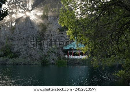 A serene gazebo with a traditional roof, nestled by a lake, surrounded by trees with sunlight filtering through