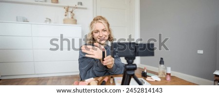 Image of young female content creator, beauty blogger sits on floor in her room, records video about makeup, shows lipstick on camera.