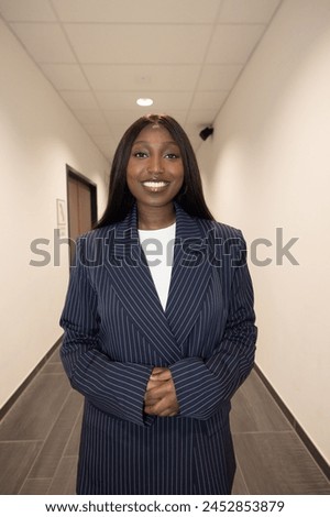 Captured in a corporate hallway, this image features a Black woman wearing a sharp pinstripe suit with a radiant smile. Her professional attire and poised demeanor convey confidence and success within Royalty-Free Stock Photo #2452853879