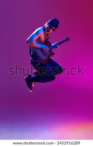 Energetic young shirtless man in sunglasses,rocket playing guitar against pink background in neon light. Concept of music, talent show, performance, concert, festival, instruments