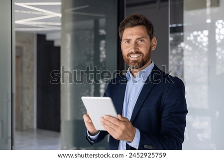 Confident and cheerful mature businessman holding a digital tablet, standing in a stylish office with a contemporary design. The natural light enhances his professional appeal.