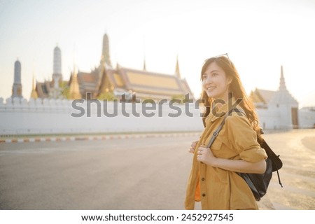 Traveler asian woman 30s, backpack slung over her shoulder, explores the intricate details of Wat Pra Kaew with childlike wonder. Sunlight dances on the golden rooftops. Royalty-Free Stock Photo #2452927545
