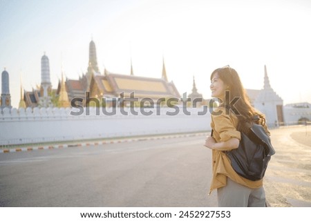 Traveler asian woman 30s, backpack slung over her shoulder, explores the intricate details of Wat Pra Kaew with childlike wonder. Sunlight dances on the golden rooftops. Royalty-Free Stock Photo #2452927553