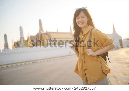 Traveler asian woman 30s, backpack slung over her shoulder, explores the intricate details of Wat Pra Kaew with childlike wonder. Sunlight dances on the golden rooftops. Royalty-Free Stock Photo #2452927557