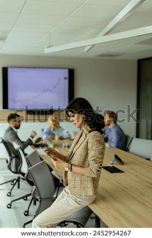 A poised woman with a tablet stands at the forefront of a collaborative office space, her team engaged in discussion behind her. Royalty-Free Stock Photo #2452954267