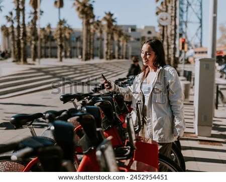 Young woman stands by a row of red bikes in sunny Barcelona, checking her phone, likely using a bike-sharing app