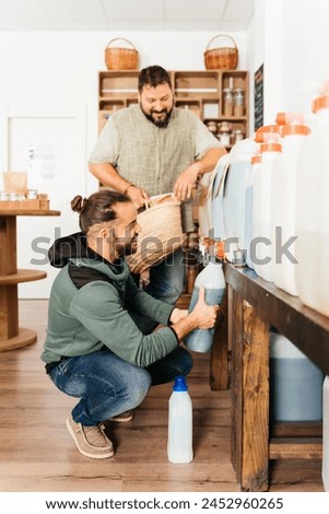 In a sustainable grocery shop, two men are refilling their bottles with liquid soap from bulk dispensers to minimize plastic use. Royalty-Free Stock Photo #2452960265