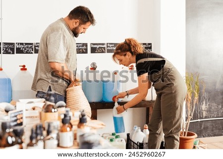 A man and woman fill containers with soap from a bulk dispenser to minimize waste in an eco-friendly shopping routine. Text: "soap, detergent, destilled water" Royalty-Free Stock Photo #2452960267